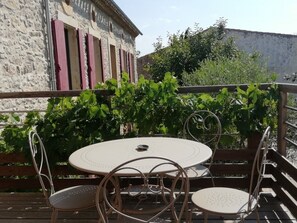 Un área de estar al aire libre con una mesa redonda y cuatro sillas, rodeada de vegetación y un edificio de piedra con contraventanas rojas.