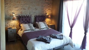A bedroom with a stone wall, a bed with a purple headboard, two bedside lamps, and a nightstand with a phone. - Au Pied du Figuier (Pauligne)