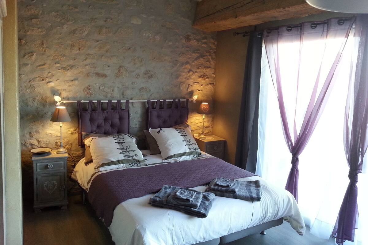 A bedroom with a stone wall, a bed with a purple headboard, two bedside lamps, and a nightstand with a phone.
