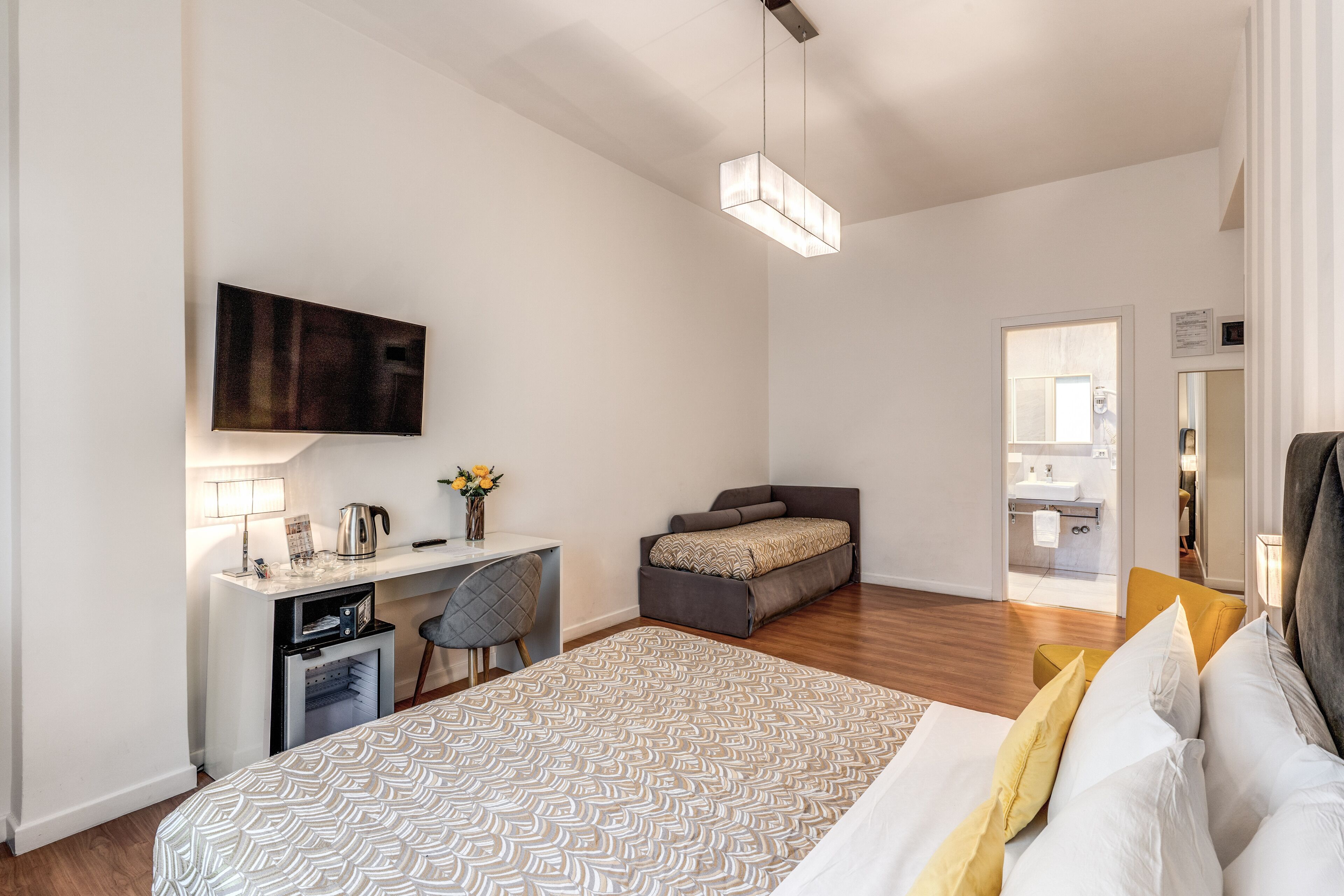 A modern bedroom with a large bed, a desk with a chair, a sofa, and a TV mounted on the wall.