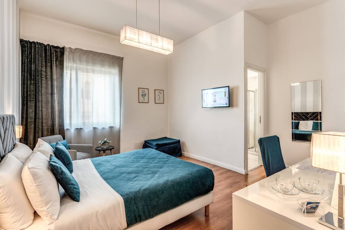 Et moderne hotelværelse med en stor seng, et skrivebord med lampe, en stol, et lille bord og et fjernsyn på væggen.