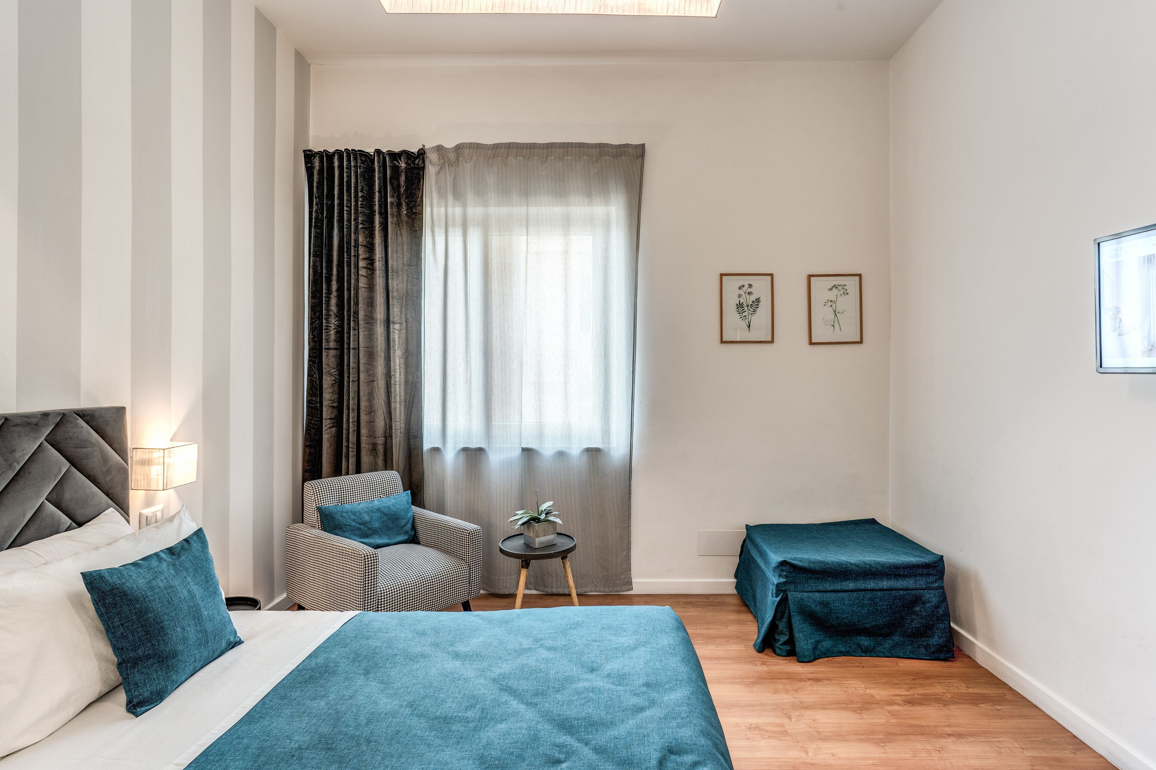 A hotel room with a bed, a chair, a small table with a plant, and a window with curtains.
