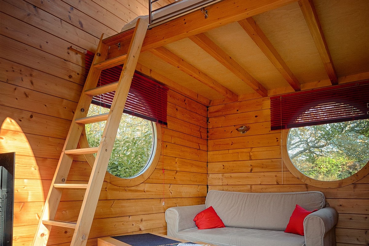 A cozy cabin interior with a loft bed, a sofa with red cushions, a wooden coffee table, and a skylight.