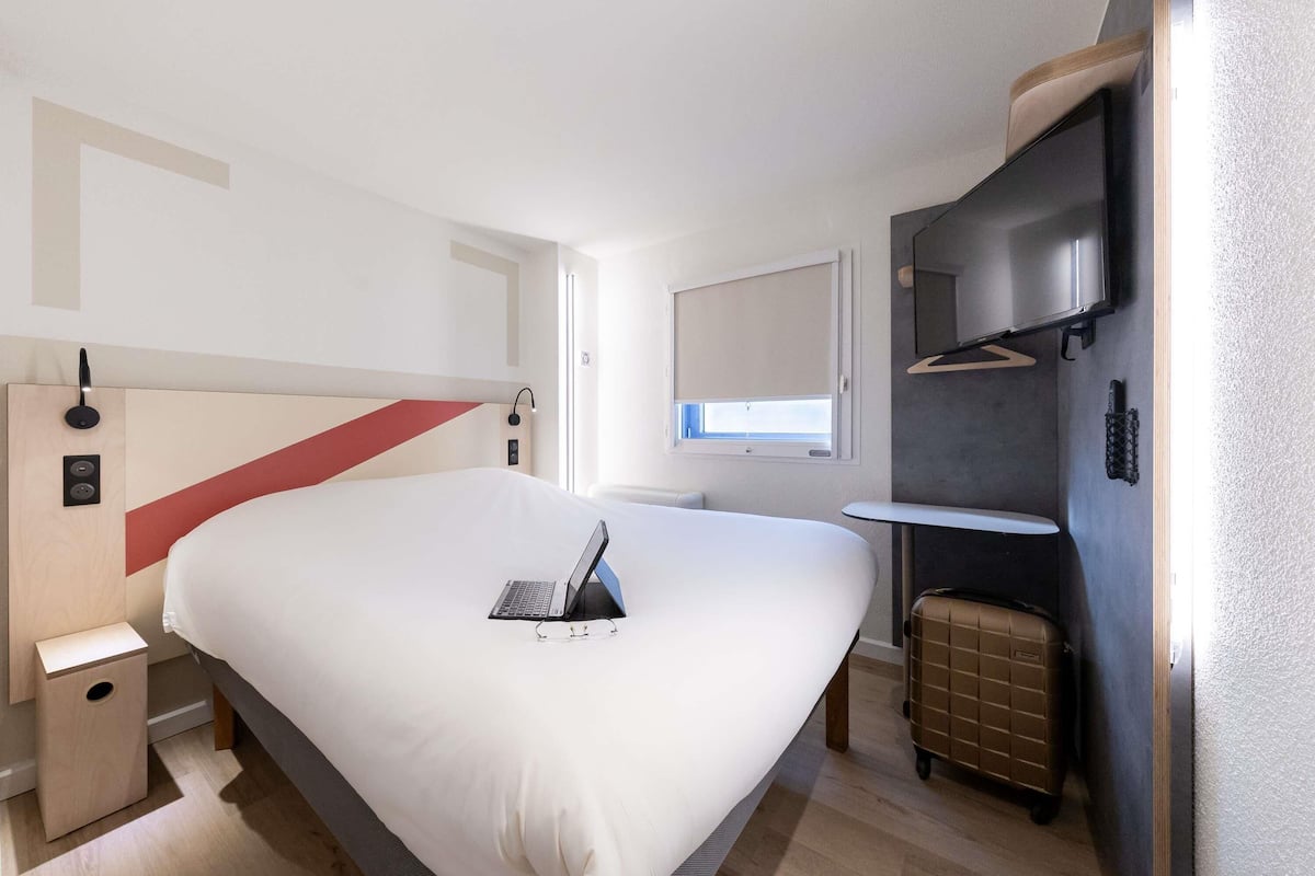 A modern hotel room with a large bed, a desk, a small table, a suitcase, and a TV.
