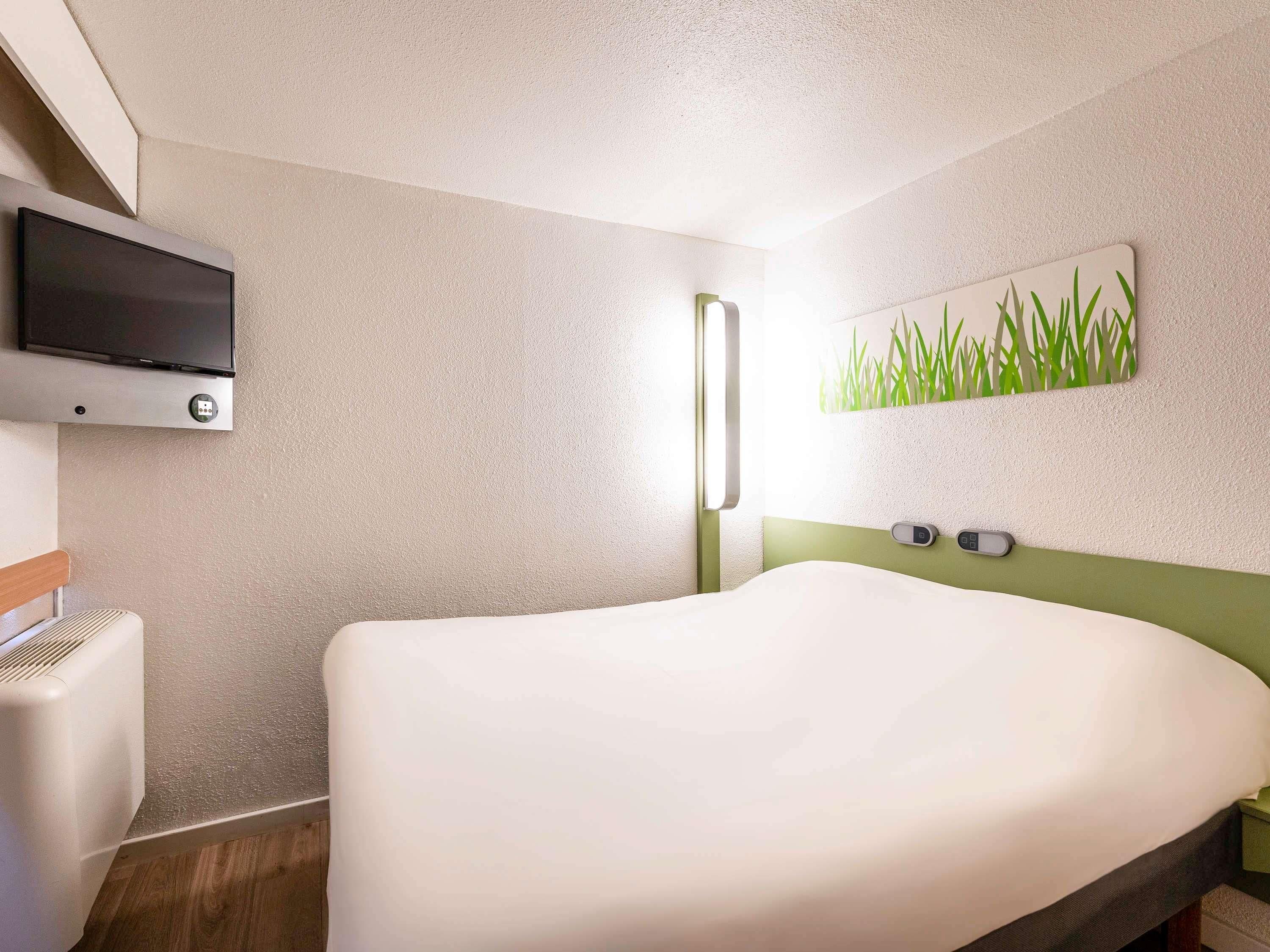 Ein Hotelzimmer mit einem Bett, einem an der Wand befestigten Fernseher, einer Wanduhr und einem Gemälde von Gras.