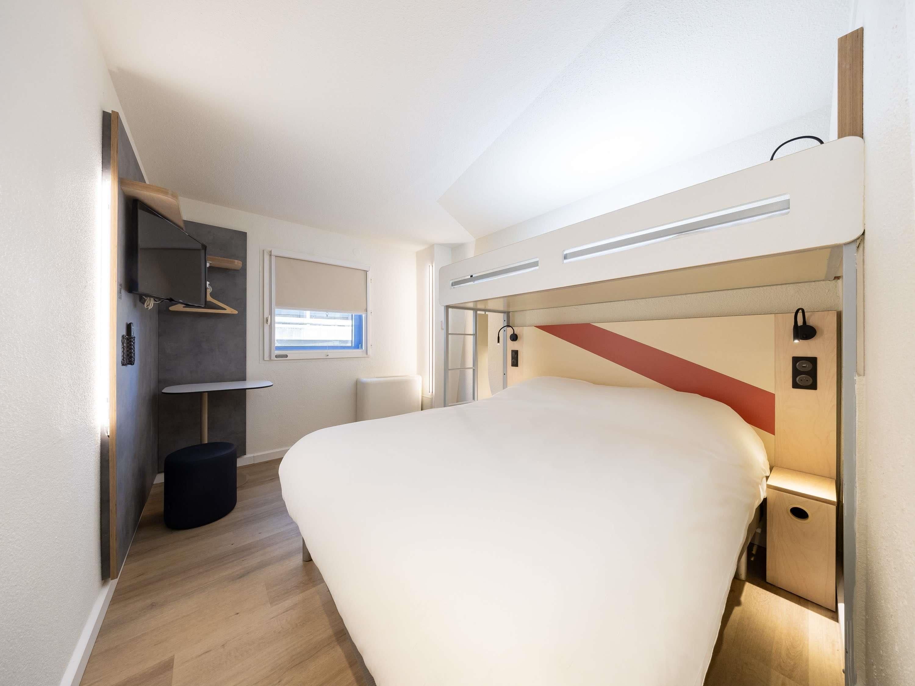 A modern bedroom with a large bed, a small desk, a chair, a wardrobe, and a window.