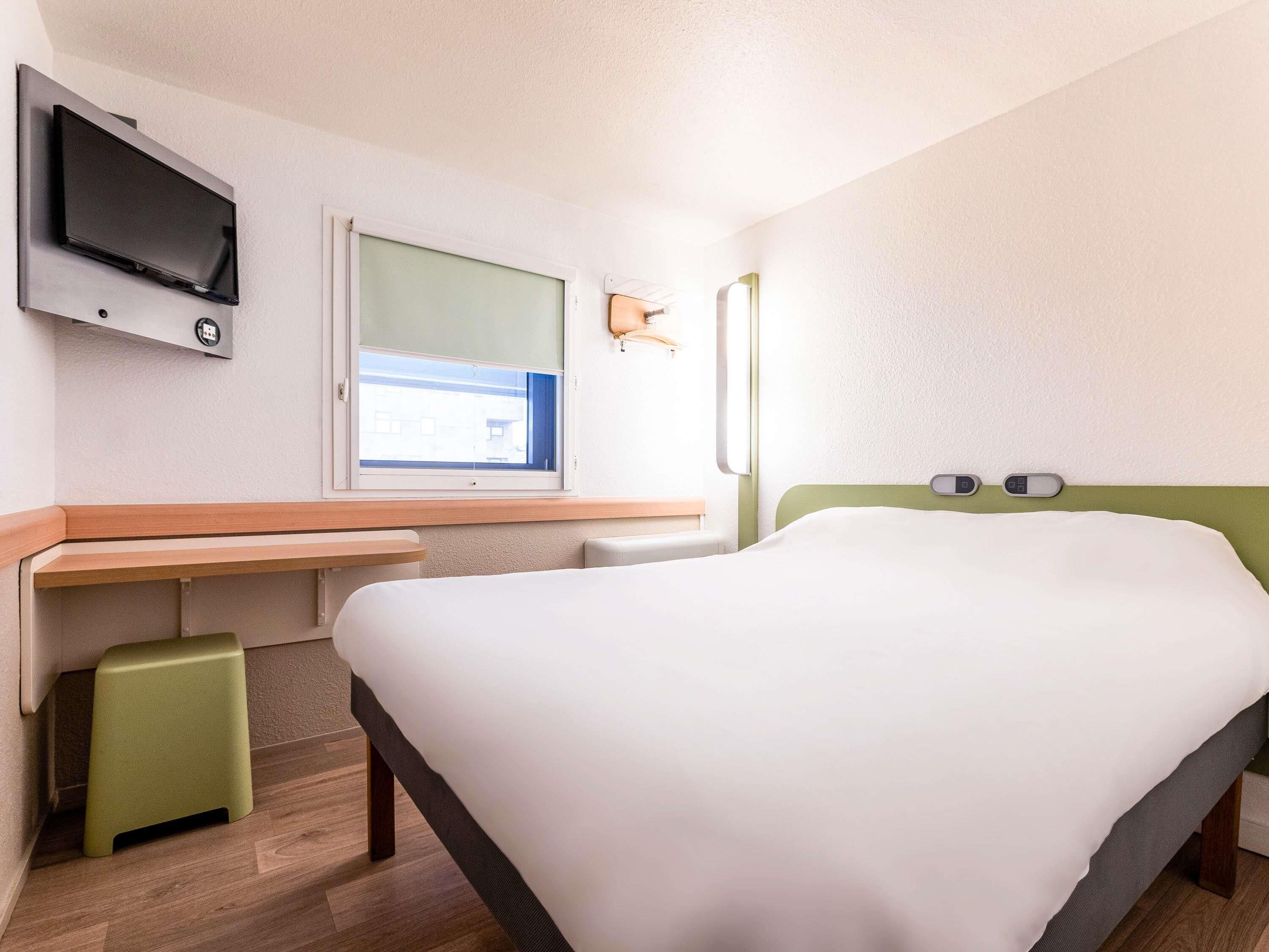 A hotel room with a bed, a TV mounted on the wall, a window, and a bedside table.