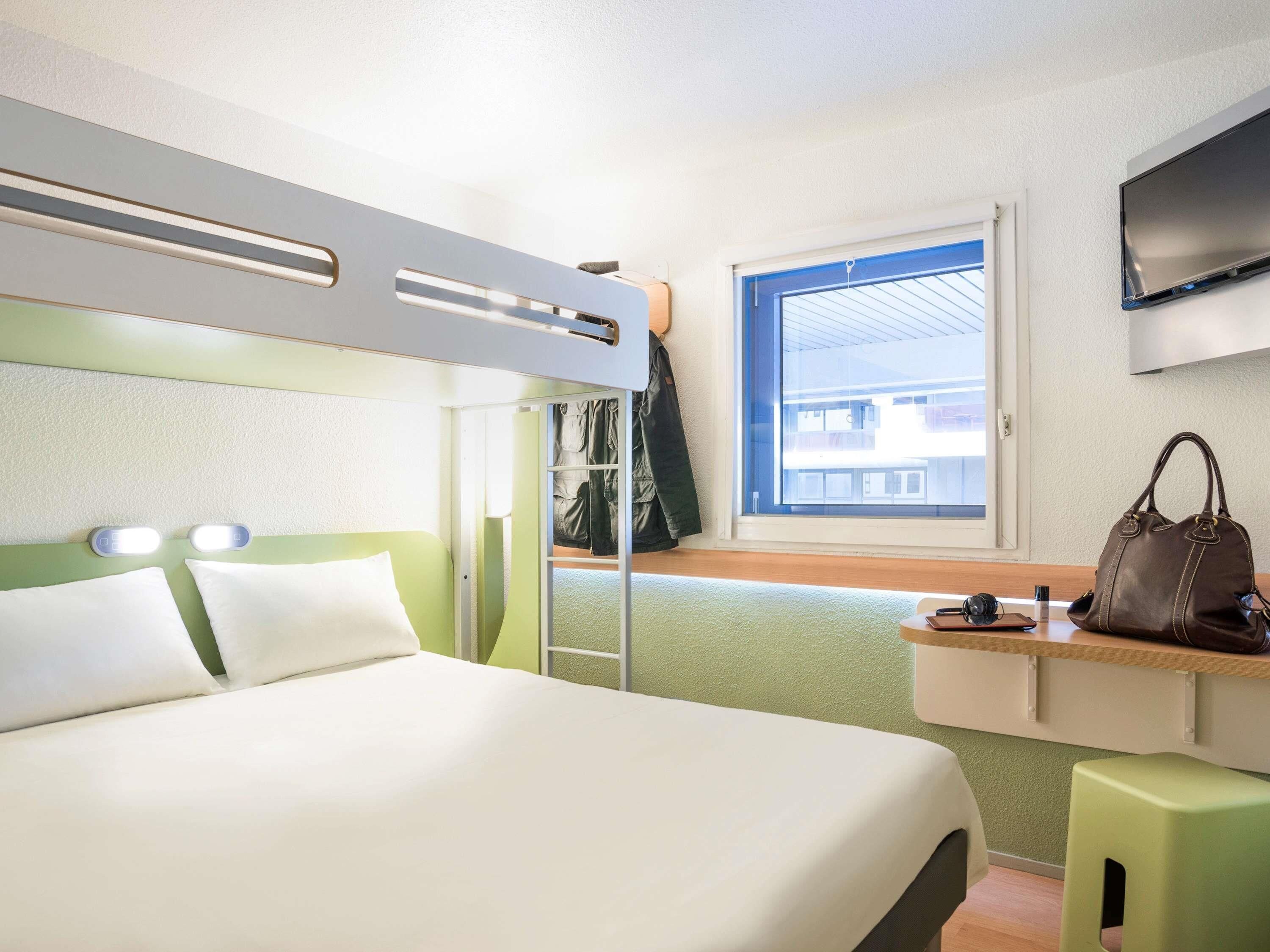 Habitación de hotel compacta con literas, un escritorio pequeño con una silla, una ventana con vistas a edificios y un televisor montado en la pared.