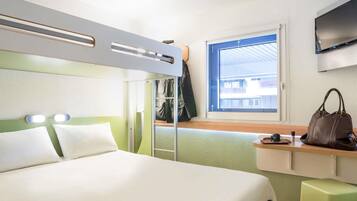 Habitación de hotel compacta con literas, un escritorio pequeño con una silla, una ventana con vistas a edificios y un televisor montado en la pared.