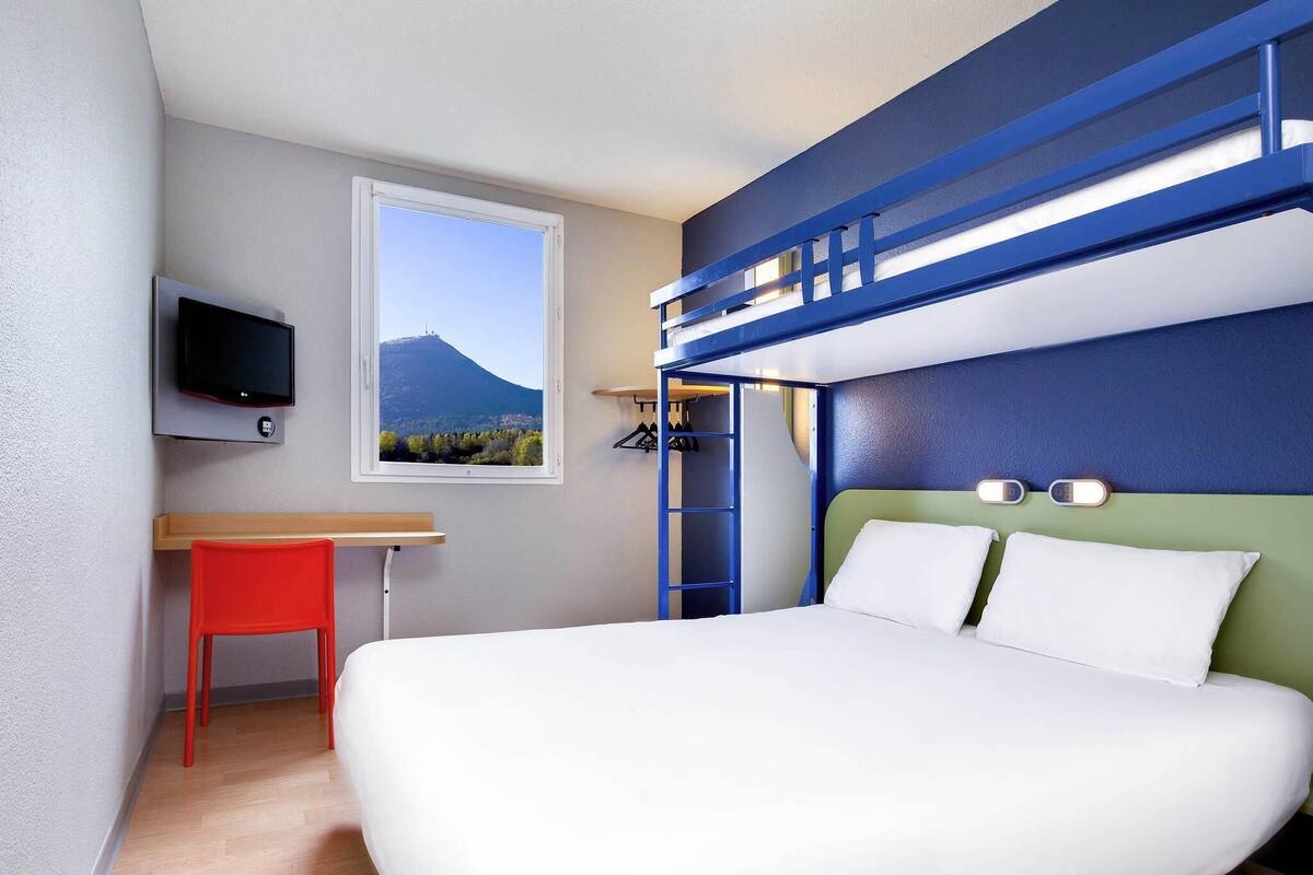 A bedroom with a bunk bed, a desk with a chair, a TV, and a window with a mountain view.