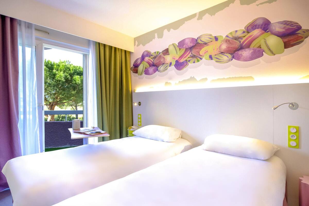 A hotel room with two beds, a window with curtains, a wall mural, a bedside table, and a green wall socket.