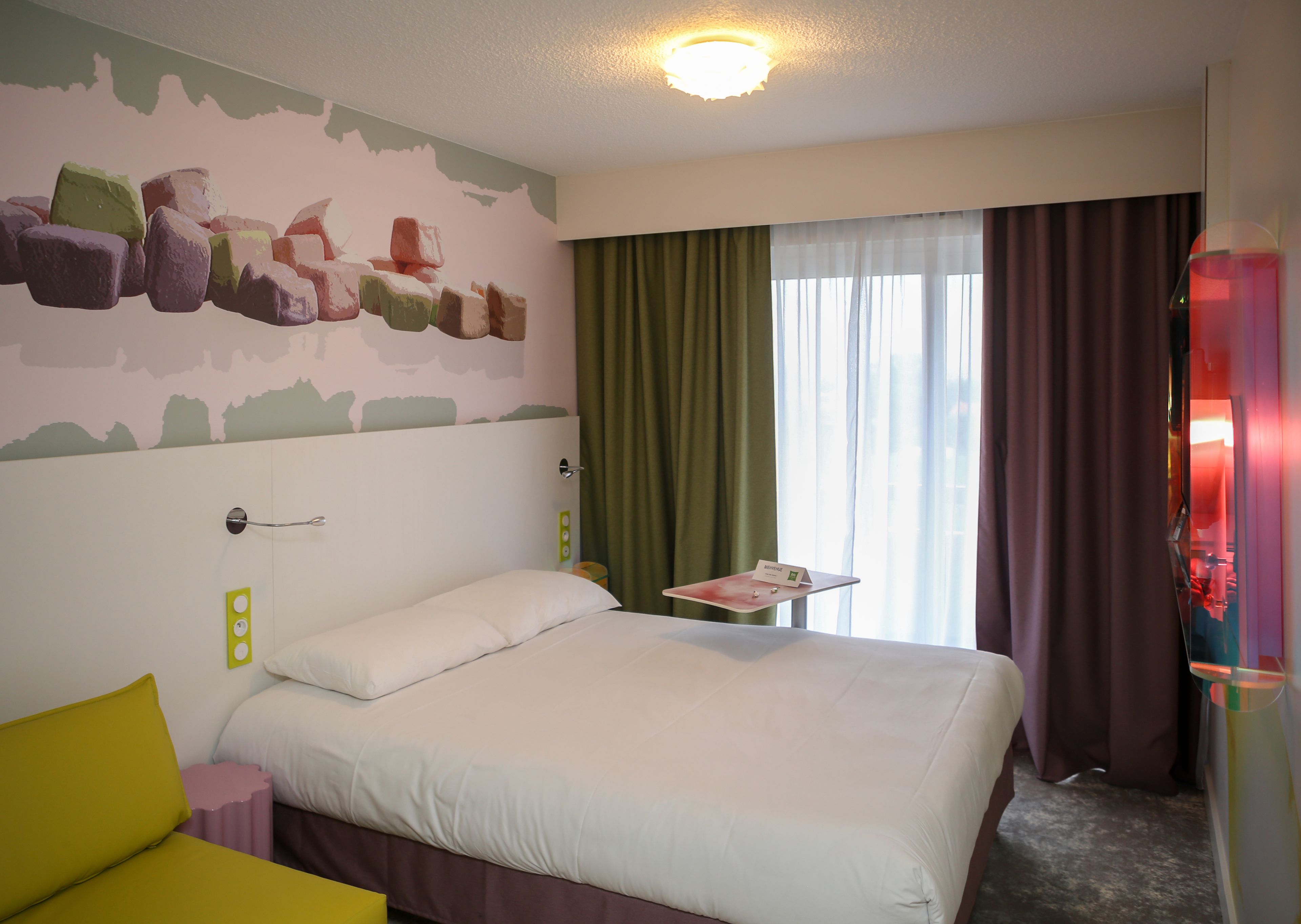 A hotel room with a bed, a small table with a name card, a green curtain, and a window with curtains.