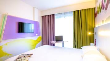 Une chambre d’hôtel avec un lit, un téléviseur fixé au mur, un ordinateur portable sur une petite table et une fenêtre avec des rideaux.