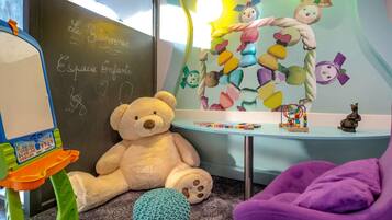 A room with a large teddy bear, a small table with toys, a chalkboard with writing, and a colorful wall mural.