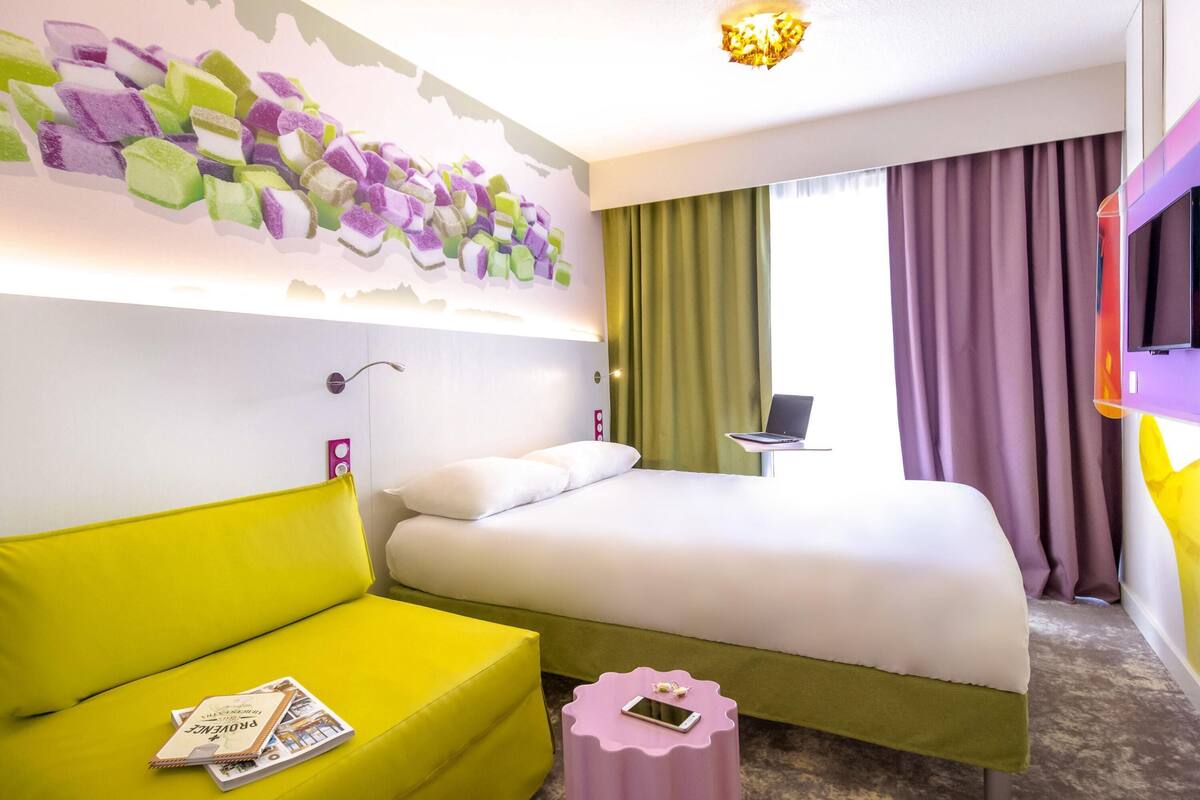 A hotel room with a bed, a yellow sofa, a small round table, a television, and a wall with a colorful abstract design.