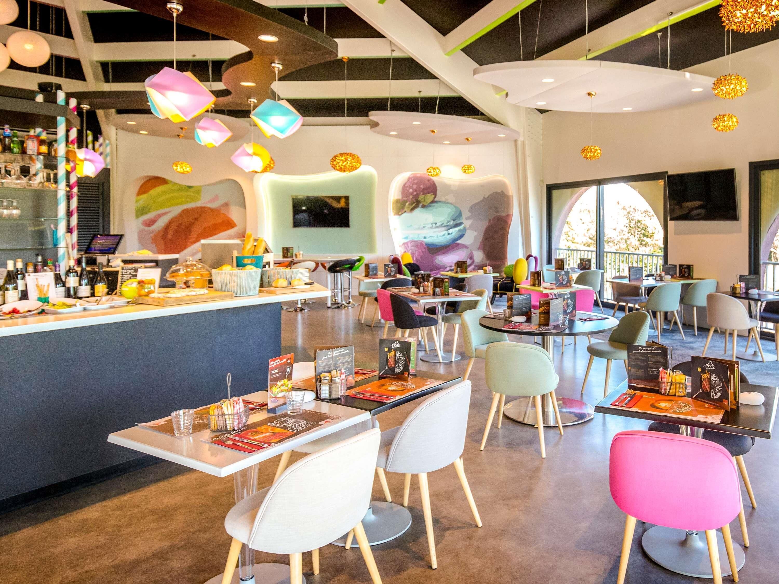 Een modern restaurantinterieur met een bar, kleurrijke hangende lampen en diverse zitmogelijkheden.