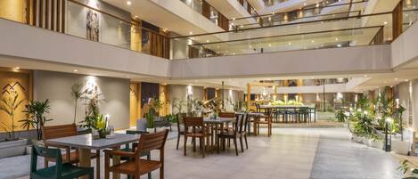A multi-level hotel lobby with glass railings, dining tables, chairs, and potted plants.