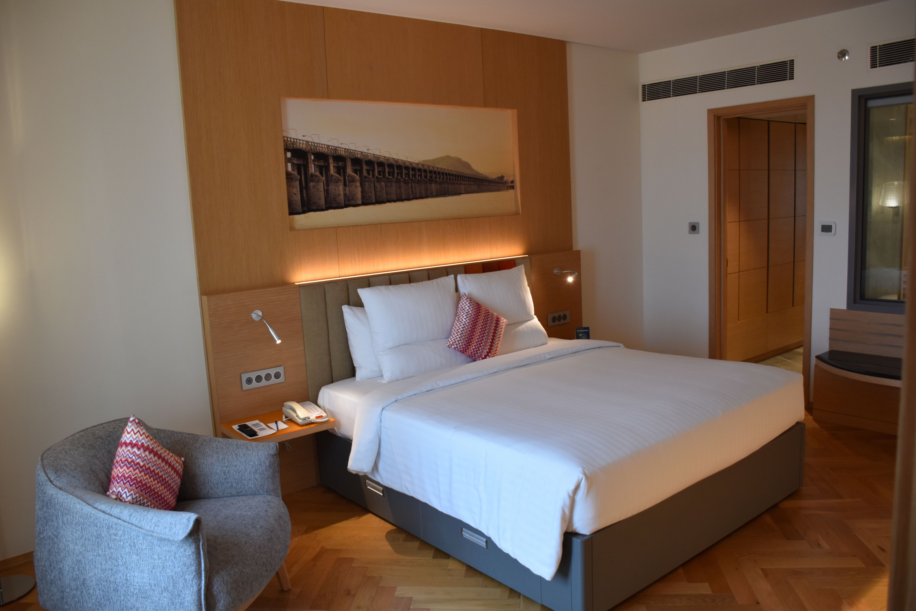 A modern hotel room with a large bed, a bedside table with a phone, a chair with a patterned cushion, and a wall-mounted artwork depicting a bridge.