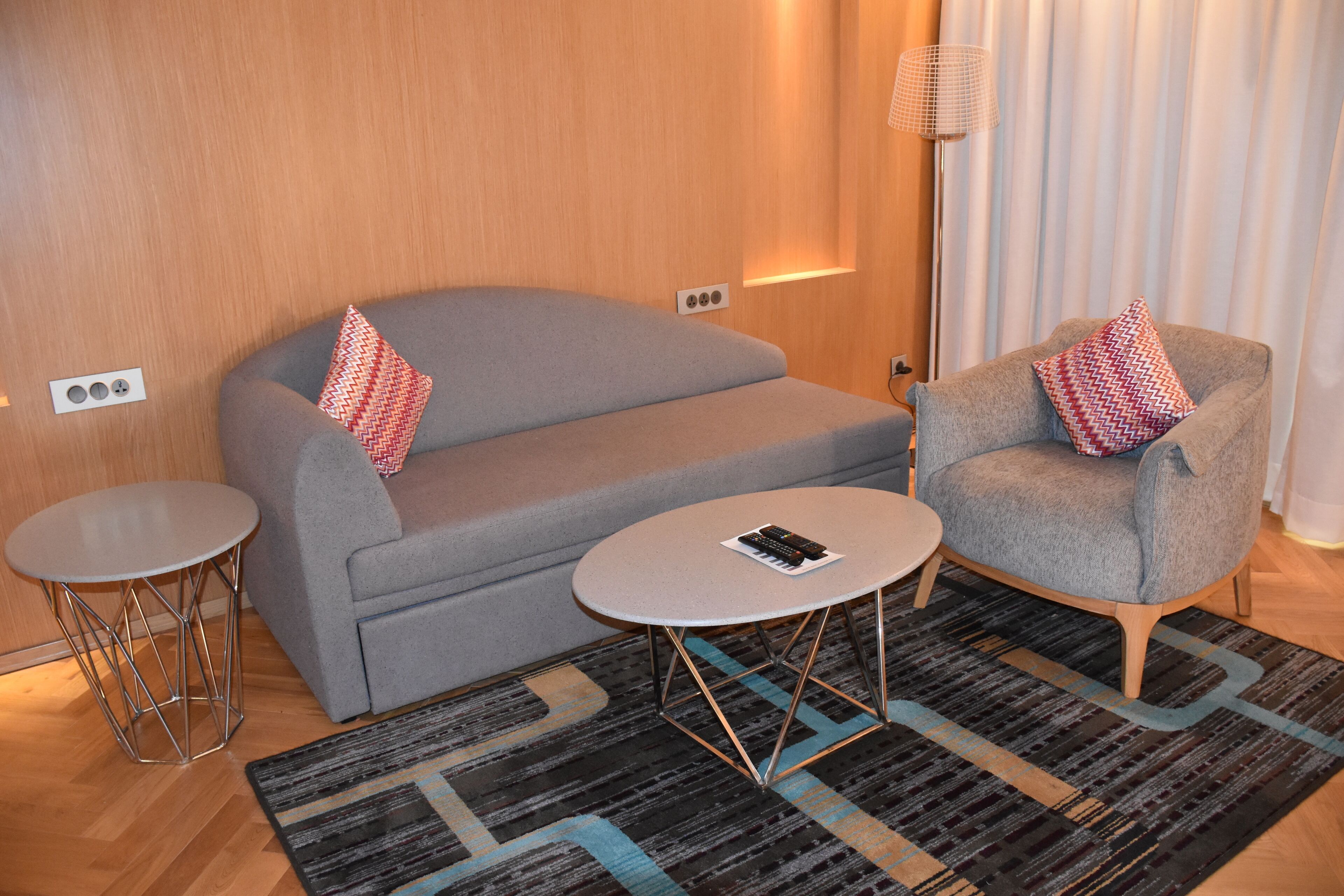 A modern hotel room with a grey sofa, a grey armchair, a small round table, and a patterned rug.