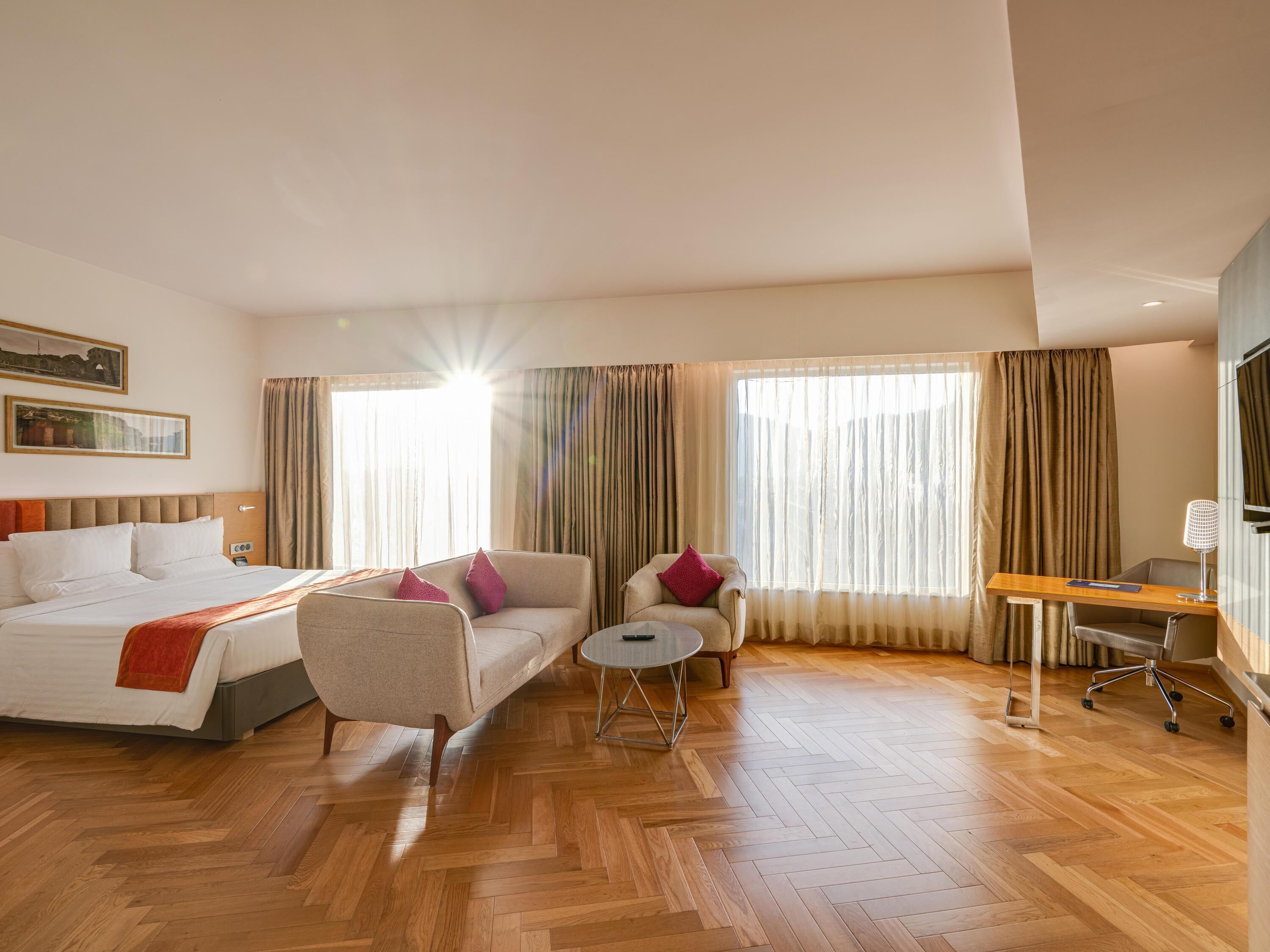 Quarto de hotel com uma cama, dois sofás, uma mesa redonda pequena, uma escrivaninha e uma televisão.