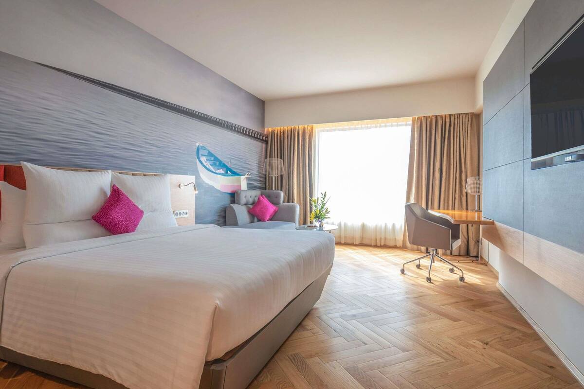 Ein Hotelzimmer mit einem großen Bett, einem Schreibtisch, einem Sessel, einem Fernseher und einem Fenster mit Vorhängen.