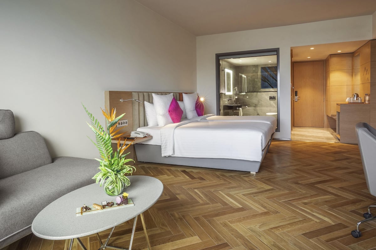 Een moderne hotelkamer met een bed, een grijze bank, een klein rond tafeltje met een plant en een houten visgraatvloer.