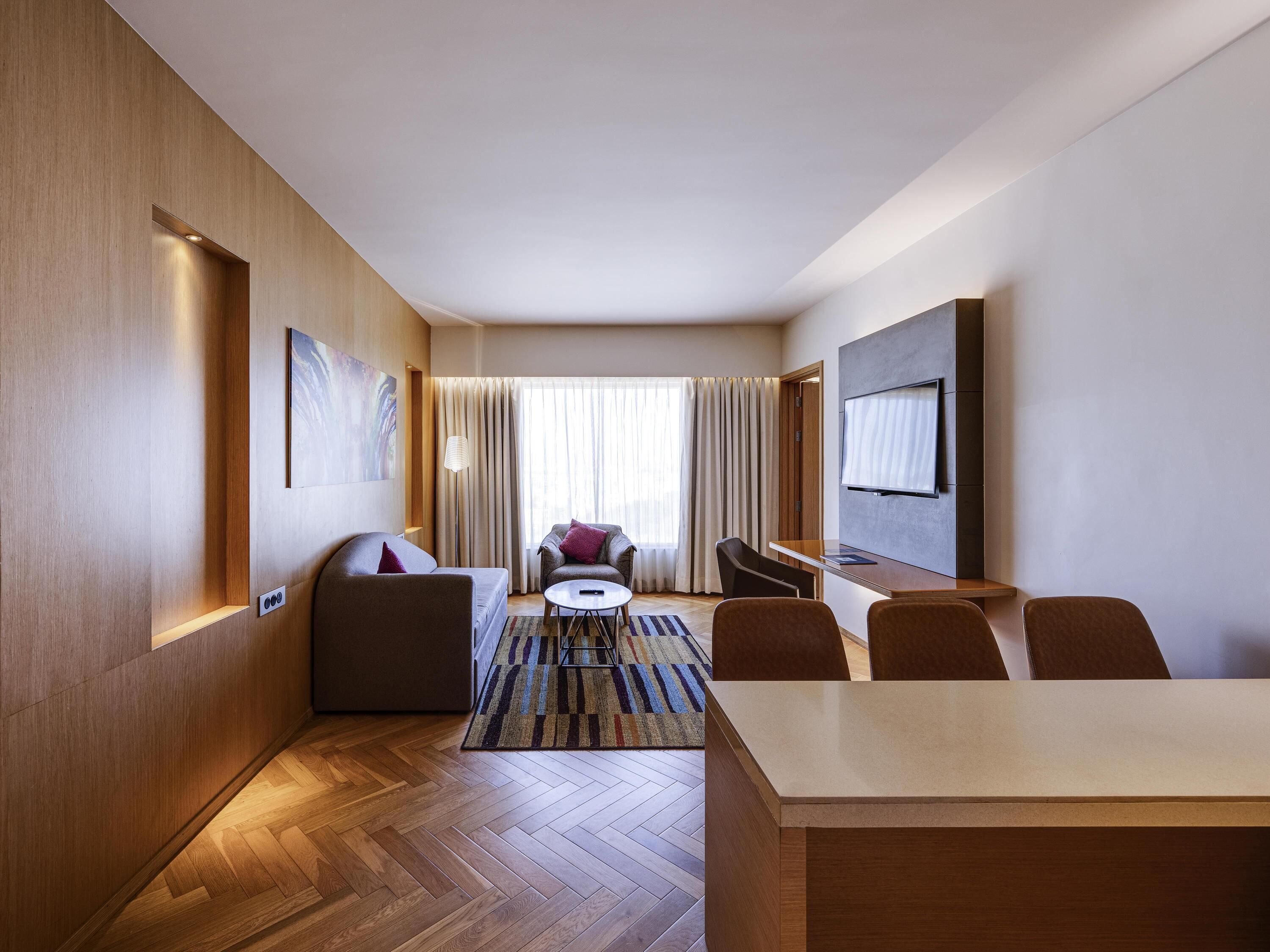 Une chambre d’hôtel moderne avec un plancher en bois, un canapé, un fauteuil, une télévision et une fenêtre avec des rideaux.