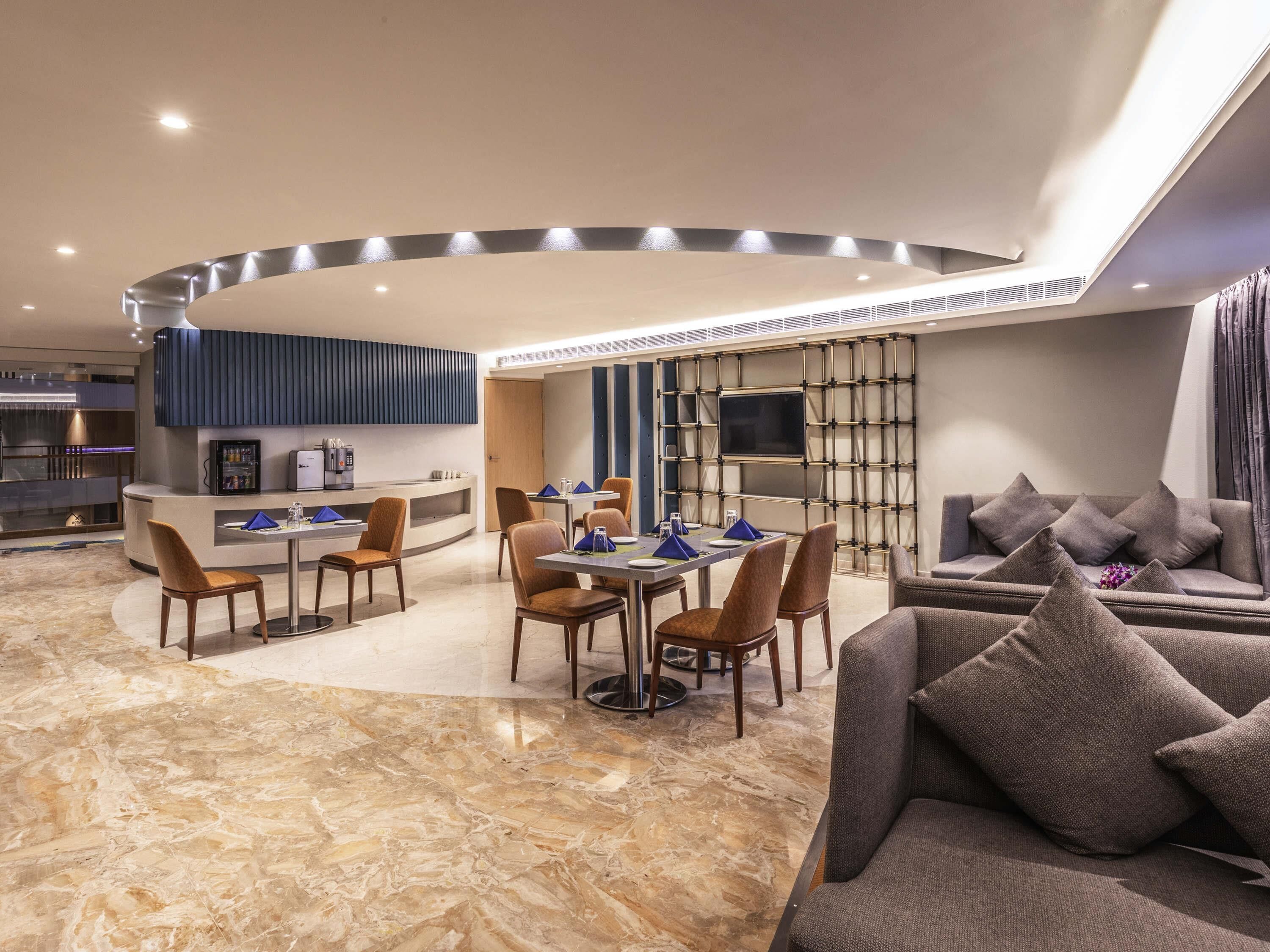 A modern hotel lobby with a dining area, a reception desk, a sofa, and a TV mounted on a wall.