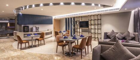 A modern hotel lobby with a dining area, a reception desk, a sofa, and a TV mounted on a wall.