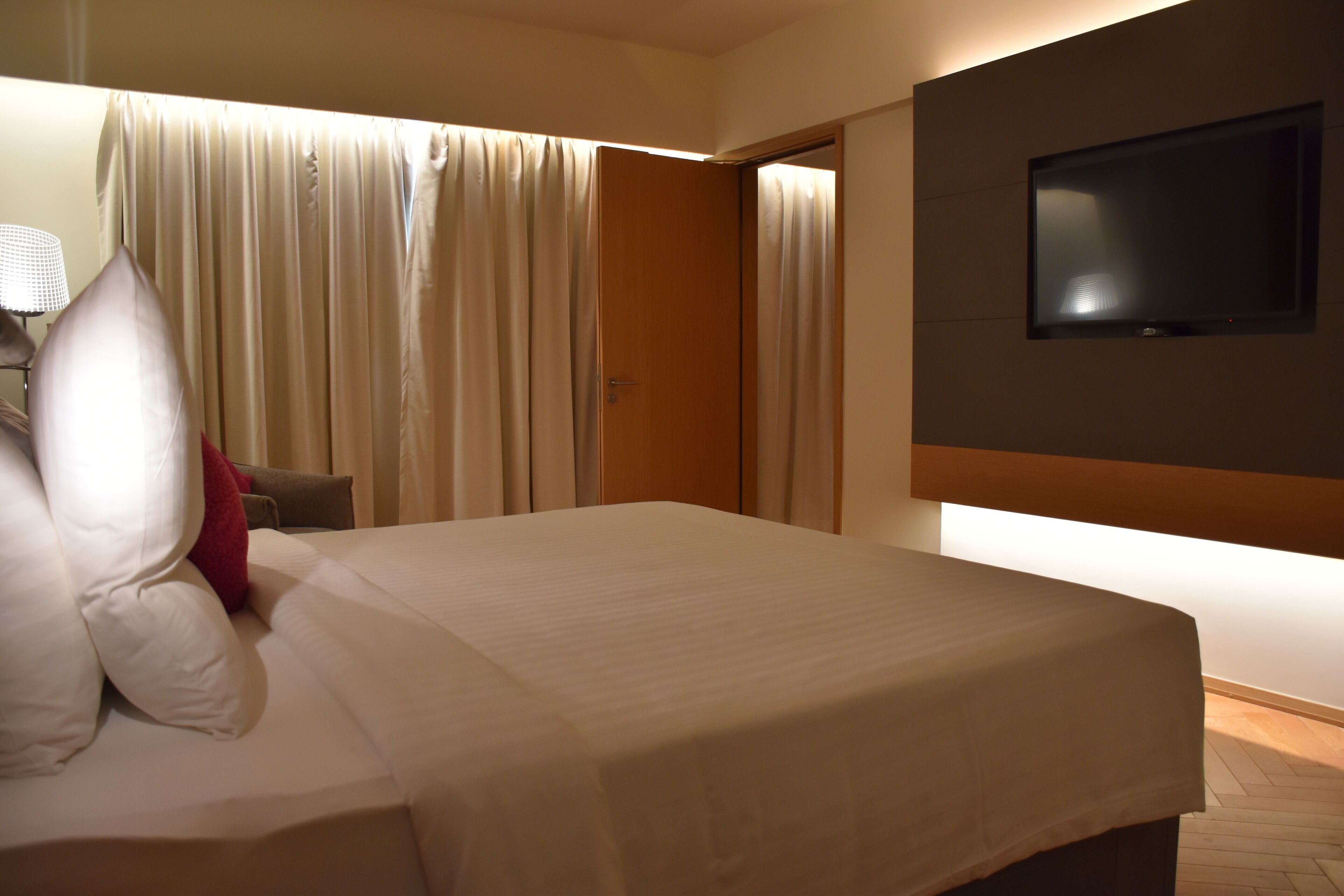 A hotel room with a bed, curtains, a TV mounted on the wall, and a lamp on a side table.