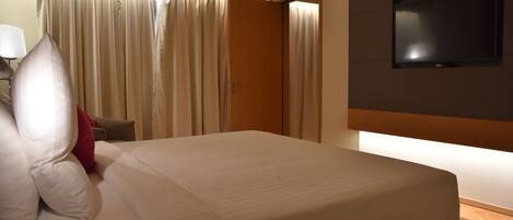 A hotel room with a bed, curtains, a TV mounted on the wall, and a lamp on a side table.