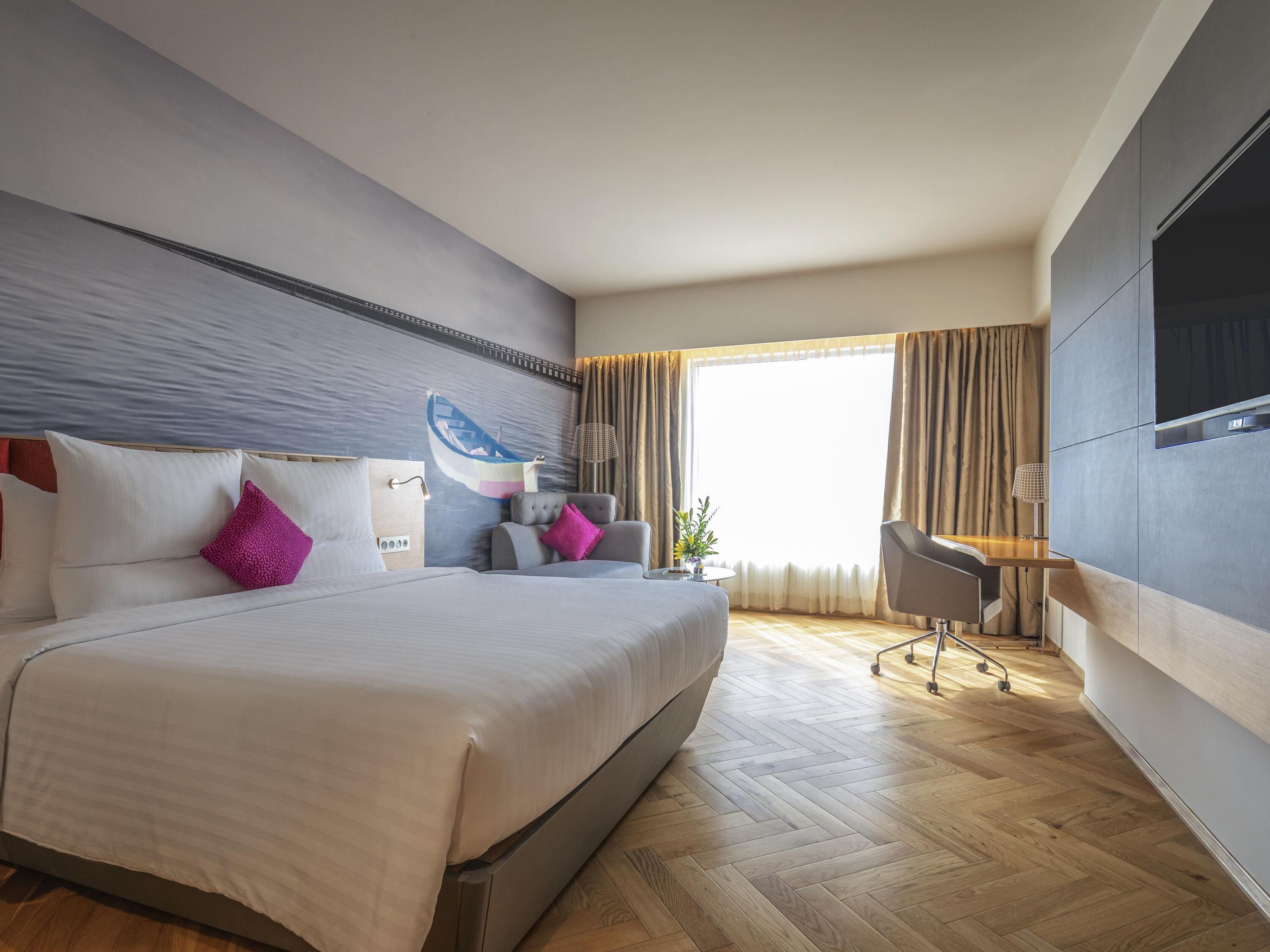 Ein modernes Hotelzimmer mit einem großen Bett, einem Schreibtisch, einem Sessel, einem Fernseher und einem Fenster mit Vorhängen.