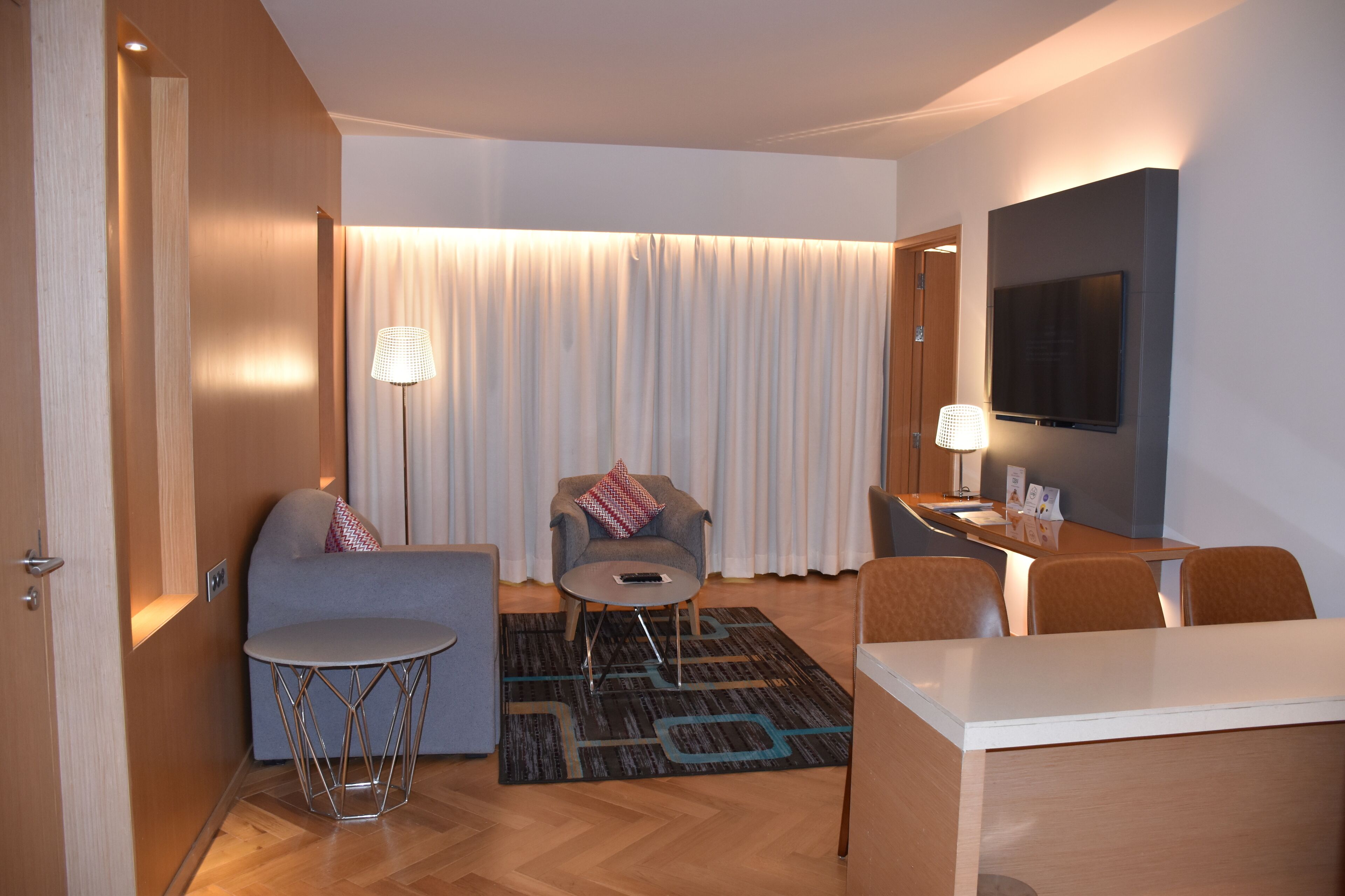 A modern hotel room with a sofa, armchair, small round table, and a TV mounted on the wall.