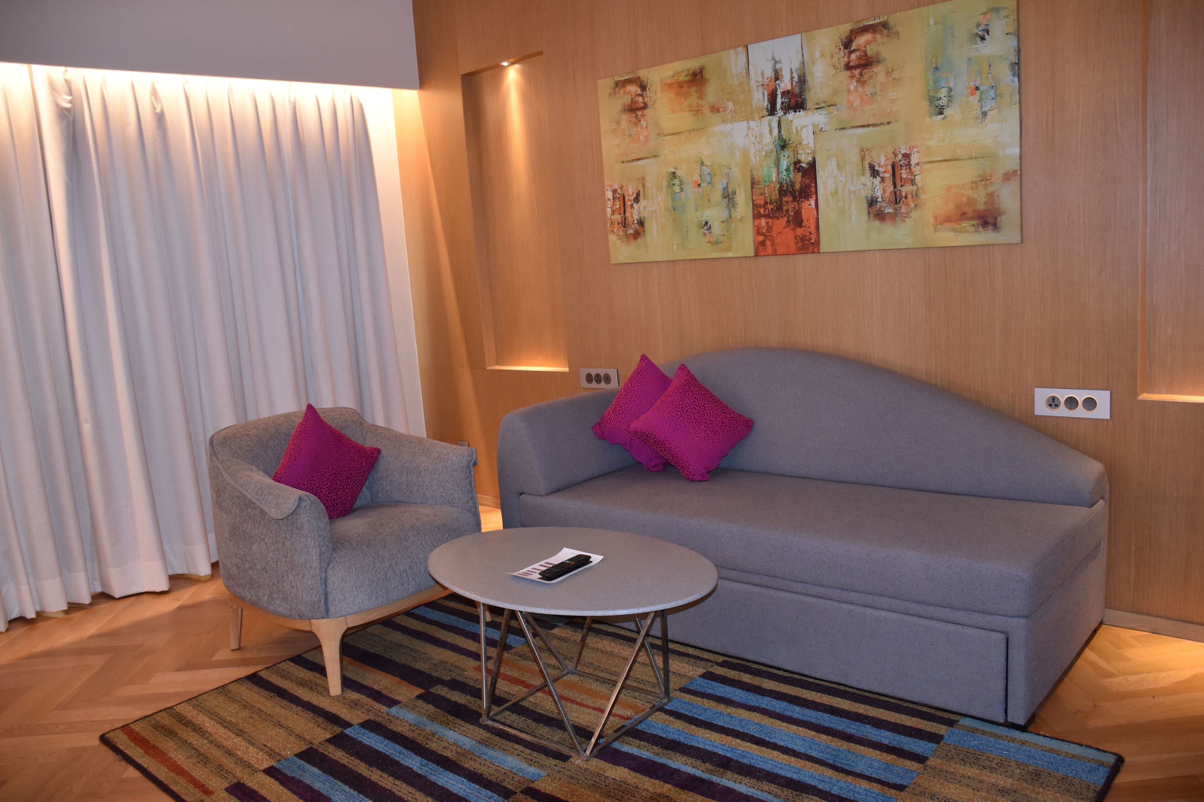 Una sala de estar moderna con un sofá gris, un sillón gris con un cojín rosa, una mesita redonda y un tapete colorido a rayas.