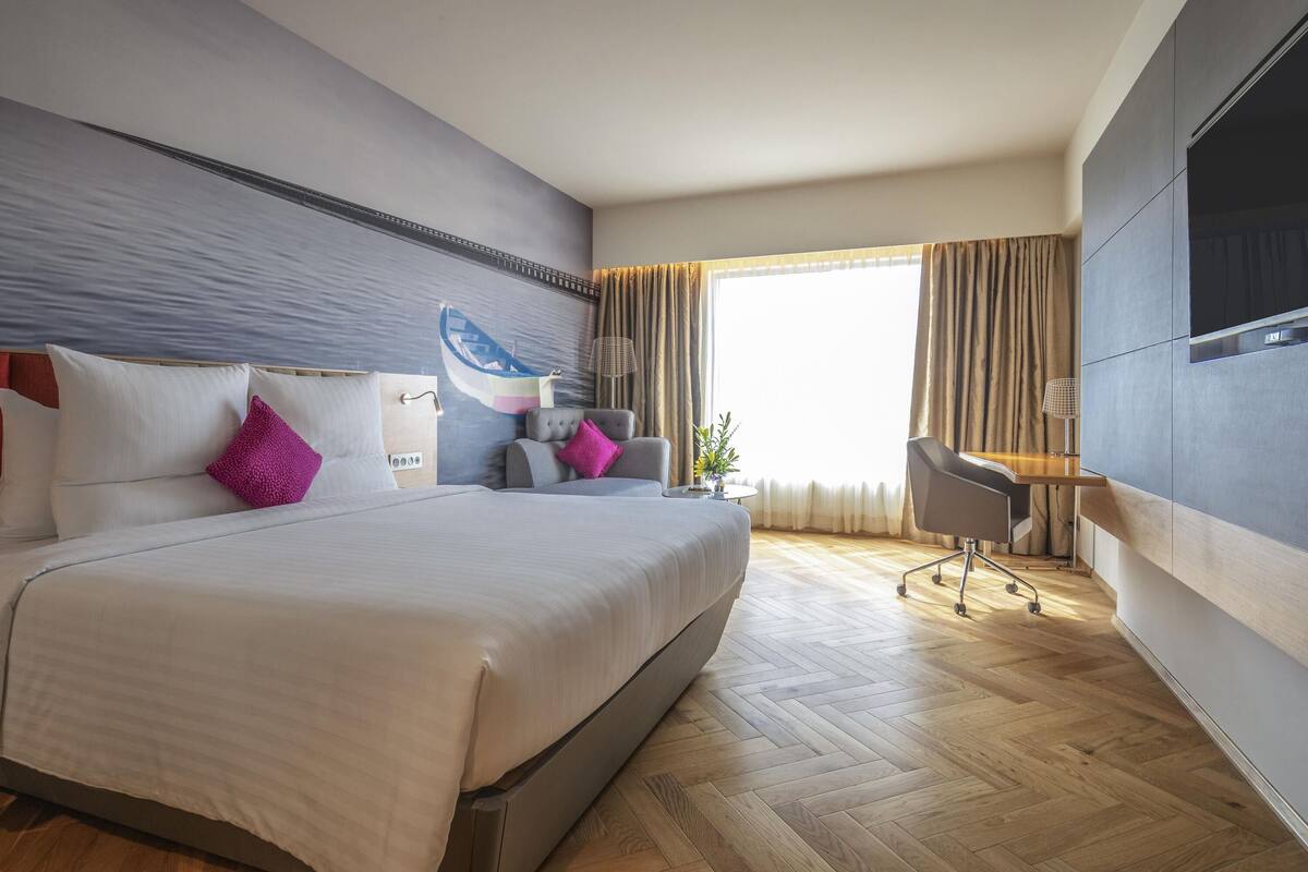 A modern hotel room with a large bed, a desk, a chair, a television, and a window with curtains.