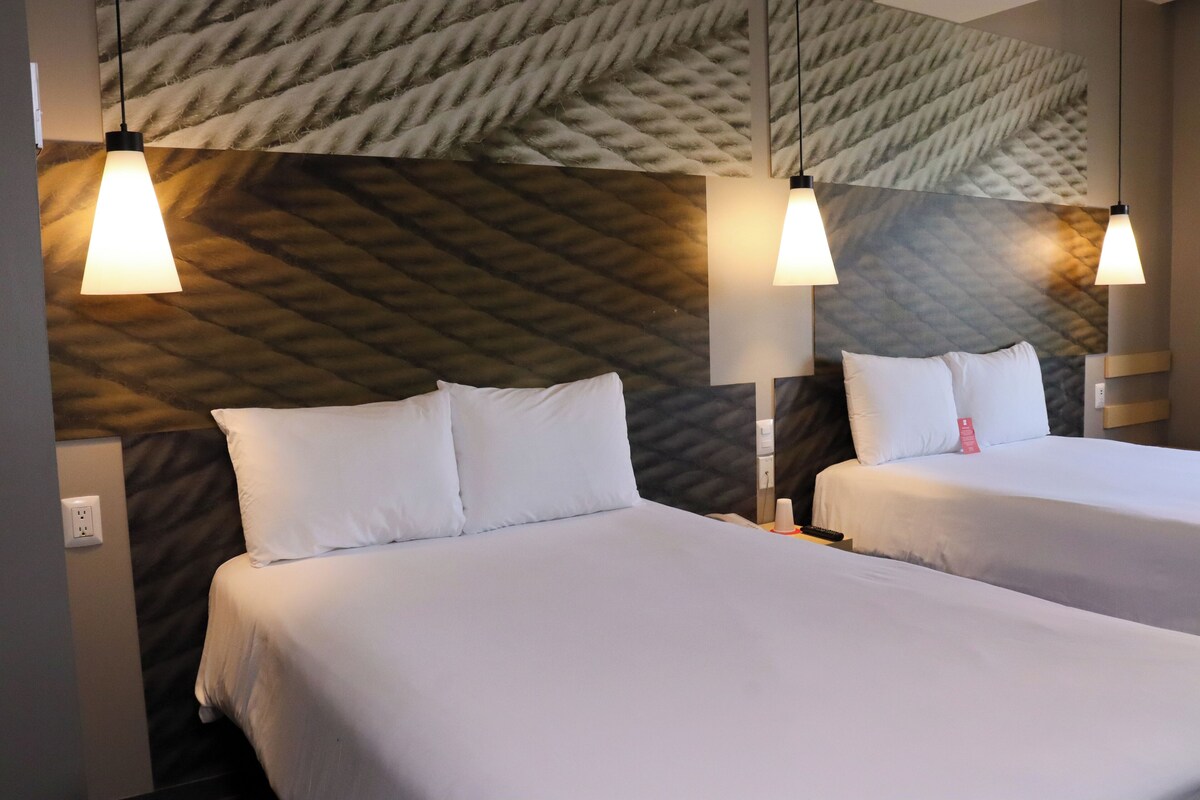 A hotel room with two beds, each with white bedding and pillows, and a textured wall behind. There are two pendant lights hanging above each bed.