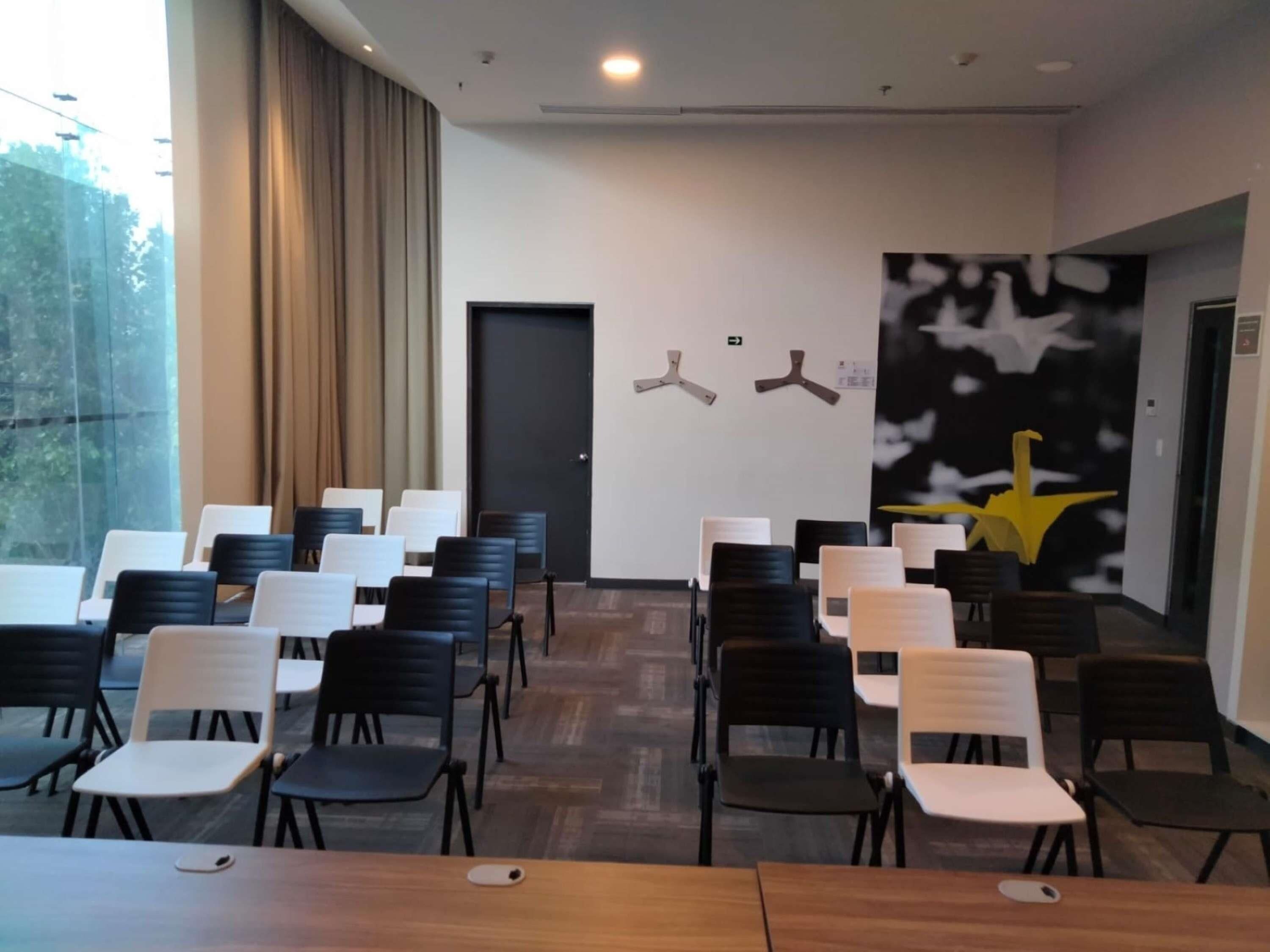 Un salón de conferencias con filas de sillas, una puerta y una pared con dos ventiladores de techo y una obra de arte abstracta de gran tamaño.