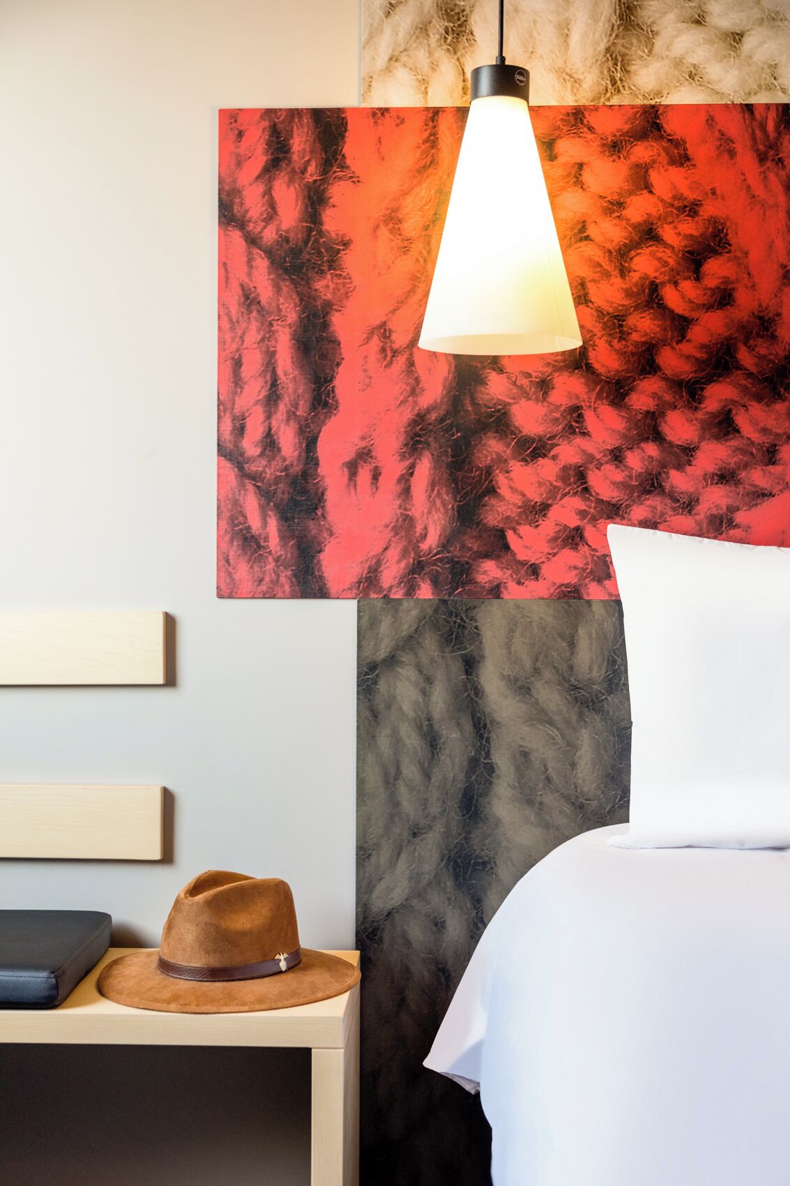 Een hotelkamer met een bed, nachtkastje, hoed en een muur met een abstract schilderij in rood.