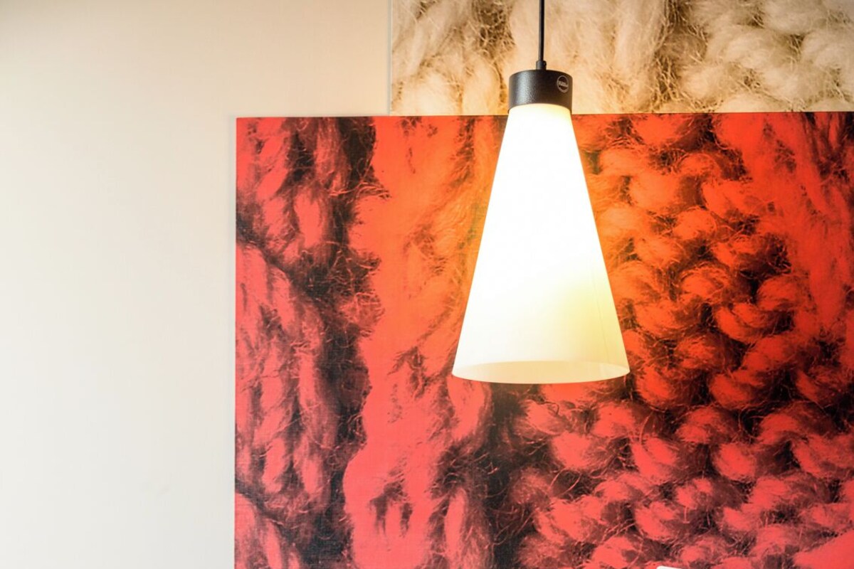 A hotel room with a bed, a nightstand, a hat, and a wall with a red abstract painting.