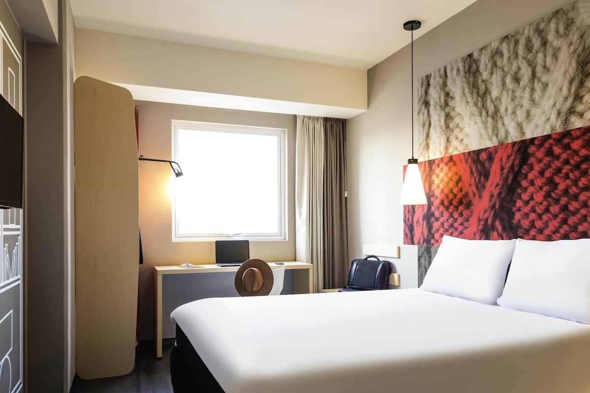Ein Hotelzimmer mit einem großen Bett, einem Schreibtisch mit einem Laptop, einem Koffer und einem Fenster mit Vorhängen.