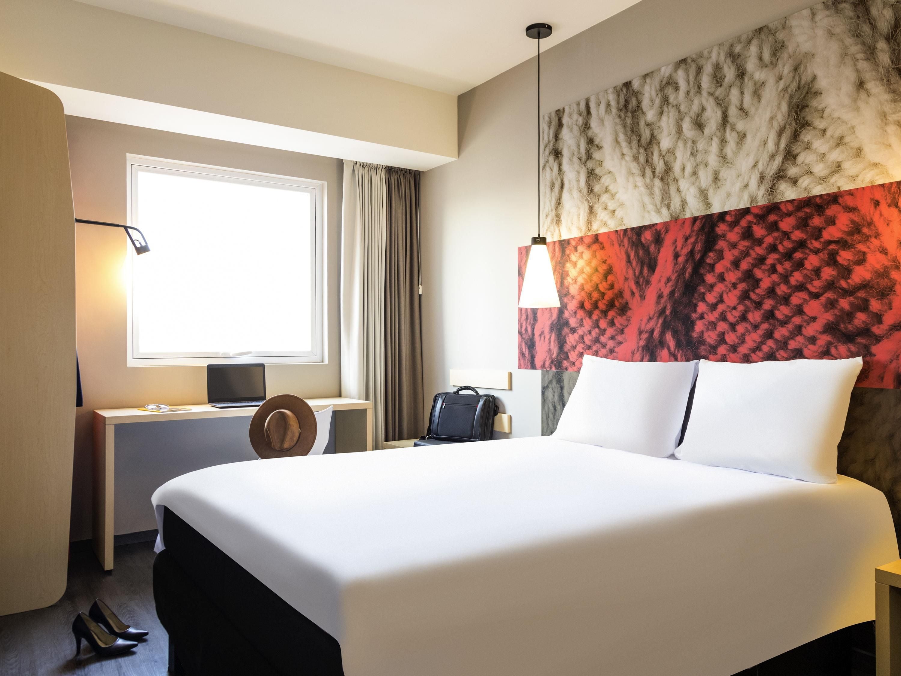 A hotel room with a large bed, a desk with a laptop, a suitcase, and a hat.