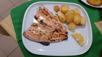 Un piatto con pesce alla griglia e patate, spicchio di limone e un fungo scuro su un piatto bianco.