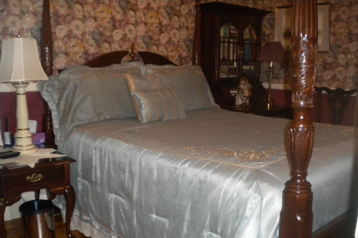 A bedroom with a four-poster bed, a nightstand with a lamp, a wooden dresser, and a floral wallpaper background.
