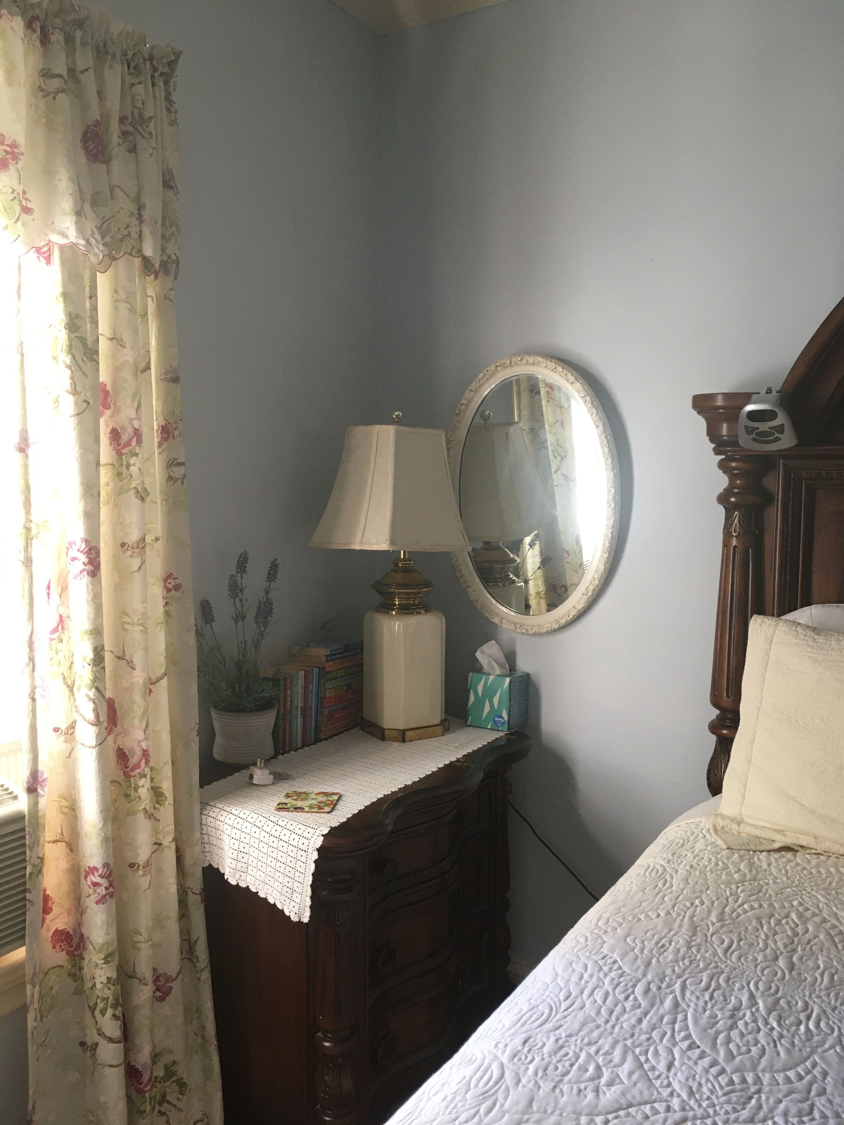 A bedroom with a bed, a dresser, a lamp, a mirror, and a window with floral curtains.