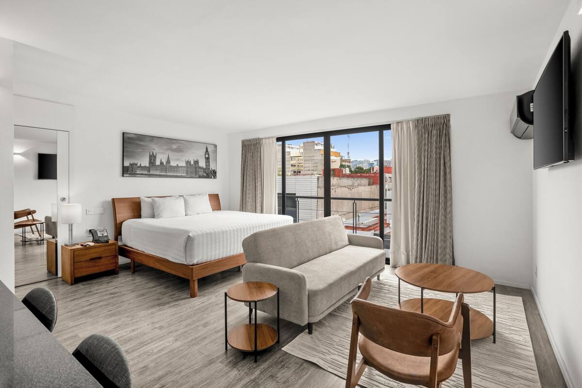A modern hotel room with a bed, sofa, two small tables, a chair, a desk, a lamp, a flat-screen TV, and a large window with a city view.