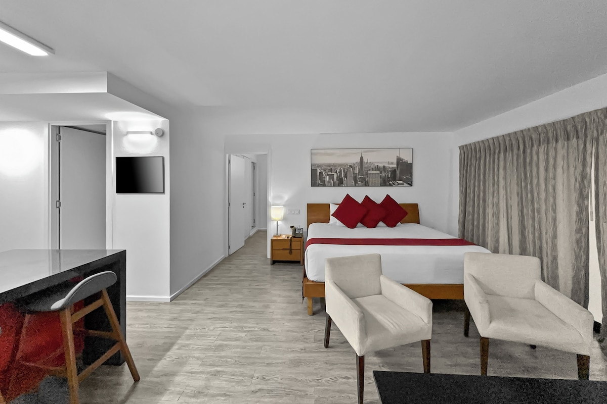 A hotel room with a bed, two chairs, a desk, a TV, a cityscape painting, and a door.