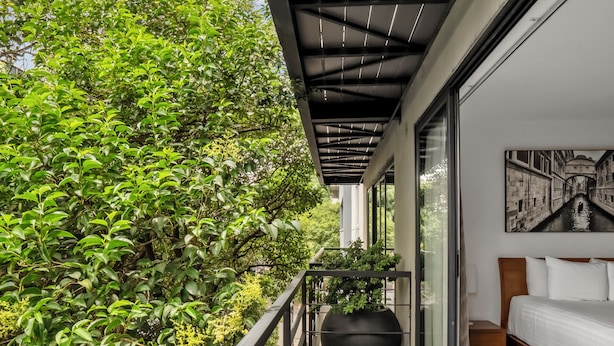A balcony with a view of greenery, a bed with white bedding, and a large black planter. - Armonik Suites (Mexico City)