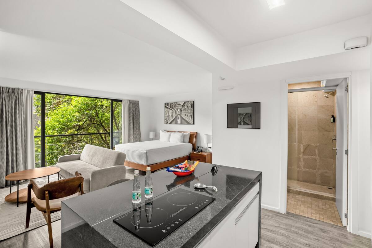 A modern hotel room with a bed, a dining area, a shower, and a view of greenery.