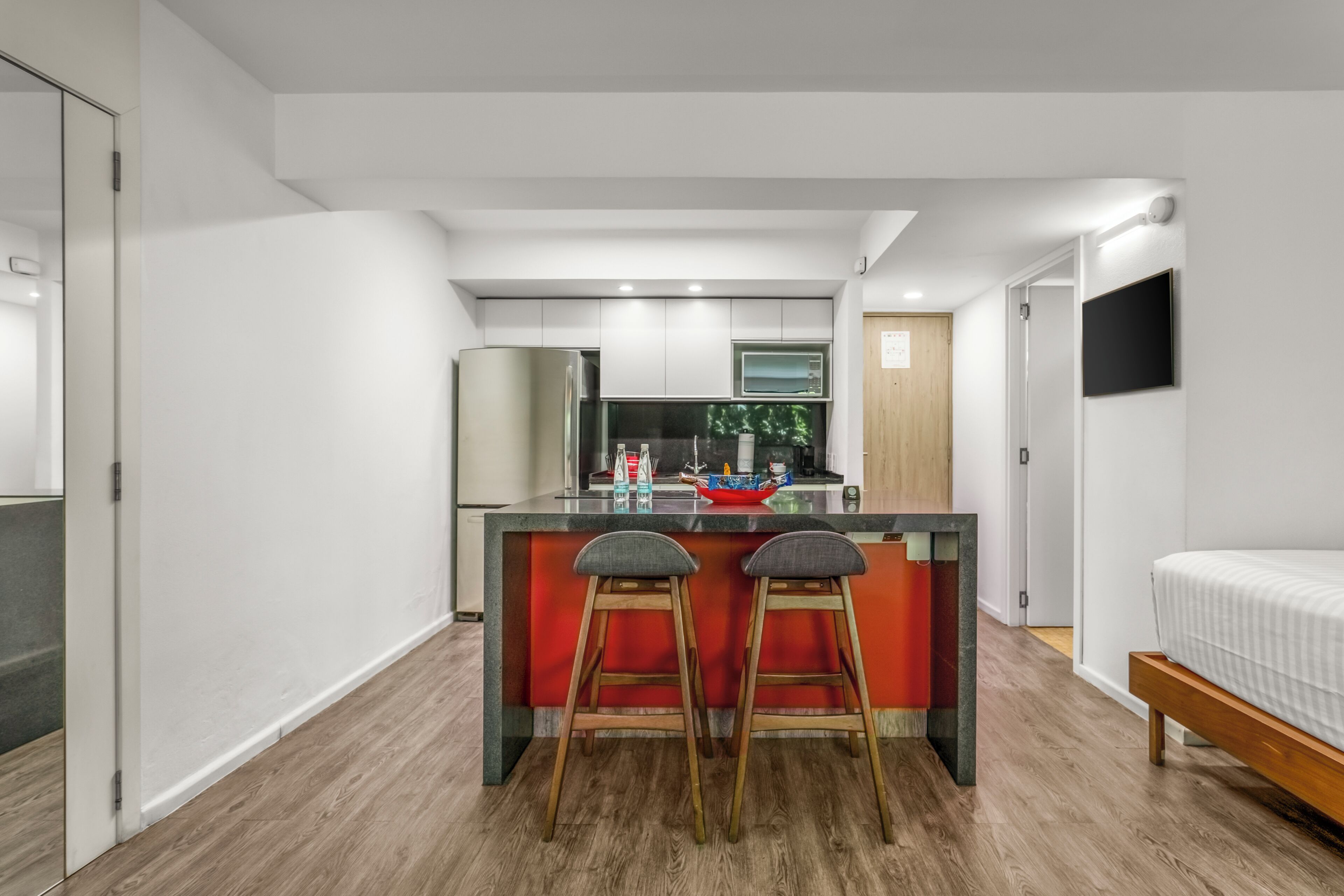 Une cuisine moderne avec un îlot central, deux tabourets de bar et un lit dans la pièce adjacente.