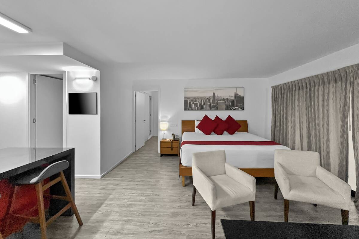 A hotel room with a bed, two chairs, a desk, a TV, a cityscape painting, and a door.